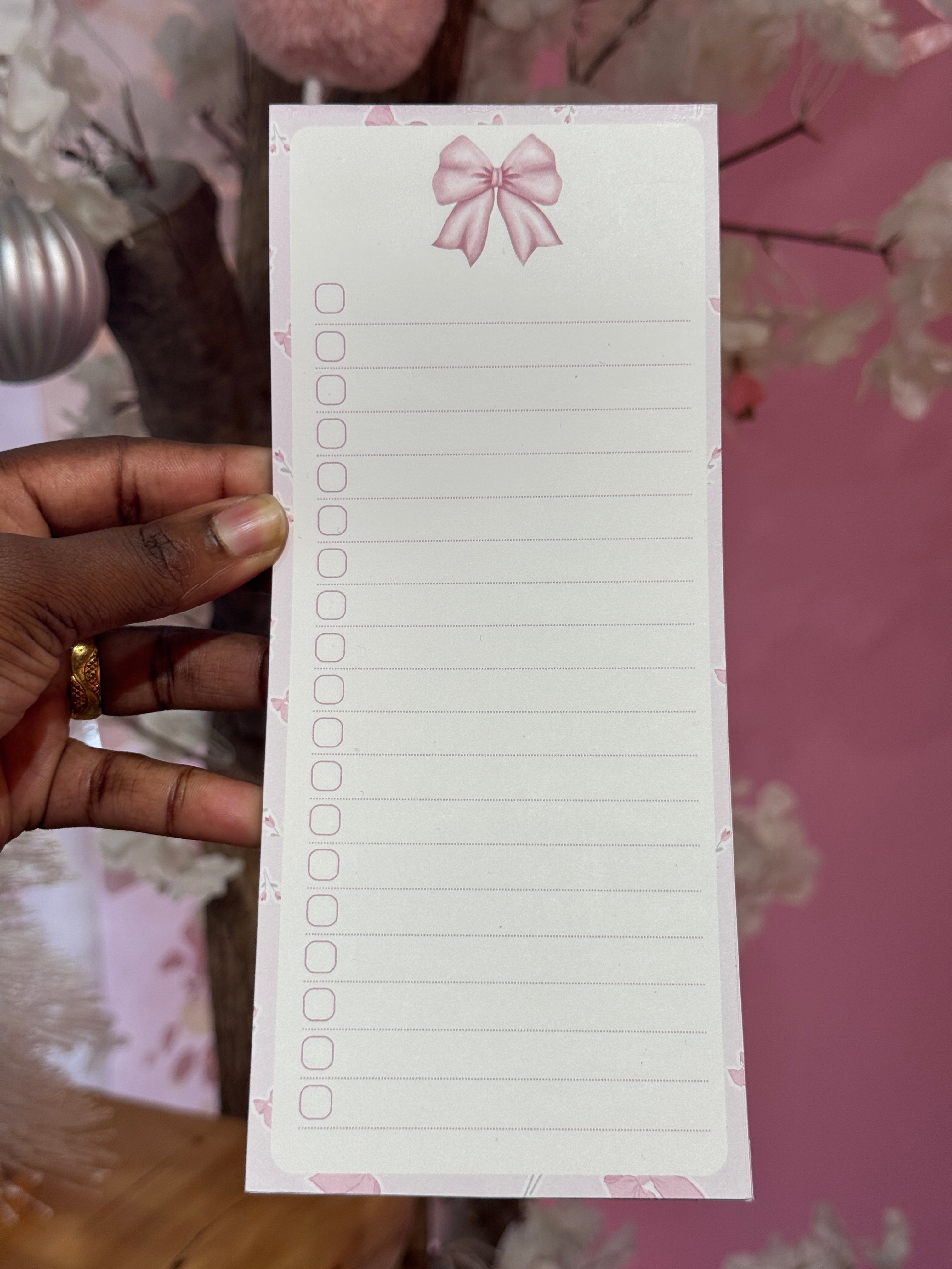 Hand holding a small notepad with a pink bow design against a floral background. pink bow. notepad. to-do list. pink to-do list