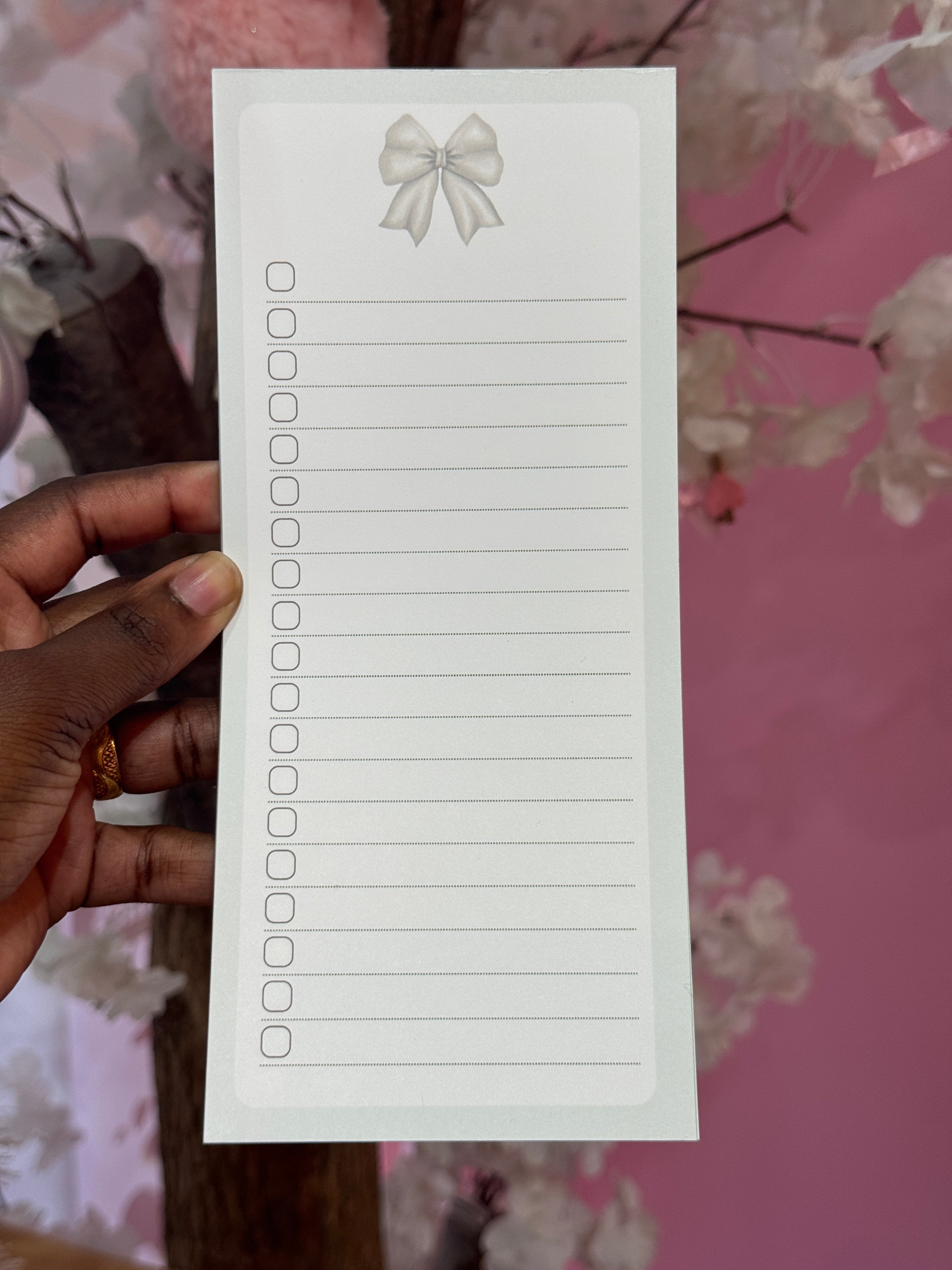Hand holding a lined notepad with decorative elements against a pink floral background. green bow. lined to do. notepad. to do list. green notepad